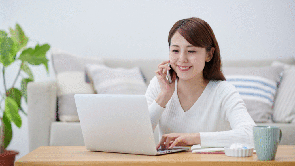 Smiling woman speaking on a smartphone while working on a laptop at home.